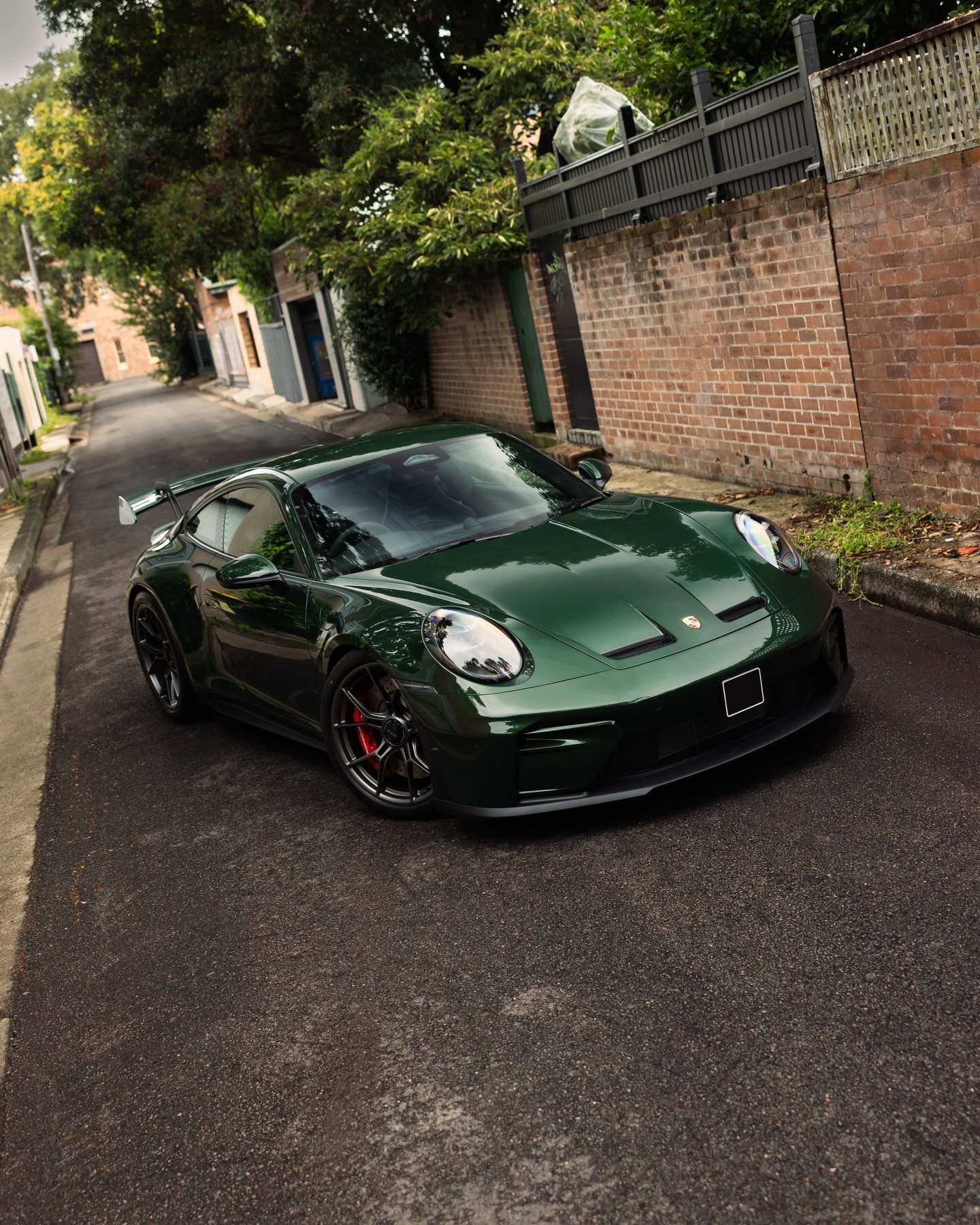 Oak Green Neo Porsche 992.2 GT3 from the front three-quarter in a lane between brick walls and trees