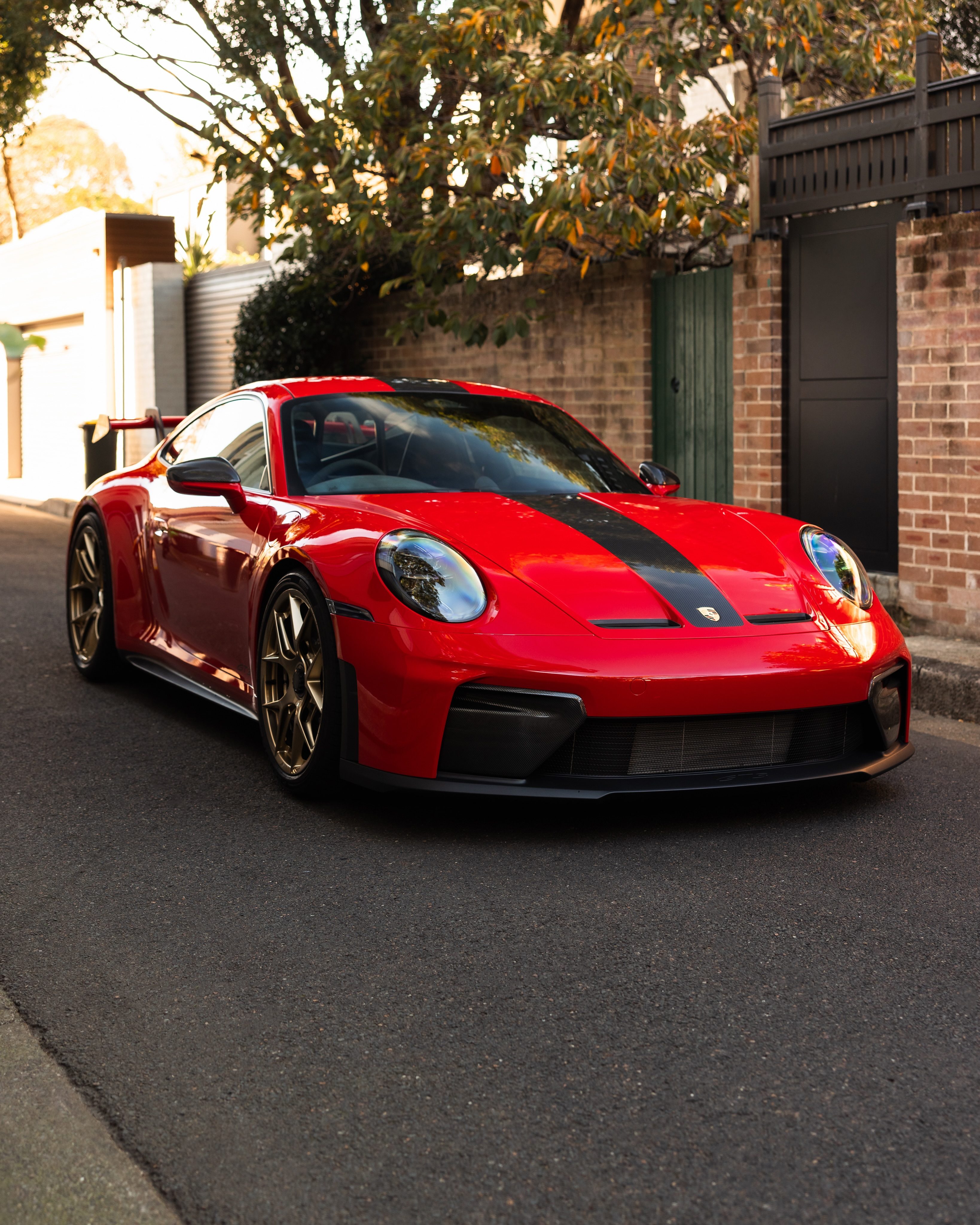 Red Porsche 992.2 GT3 with a black stripe and gold wheels in a tree-lined lane