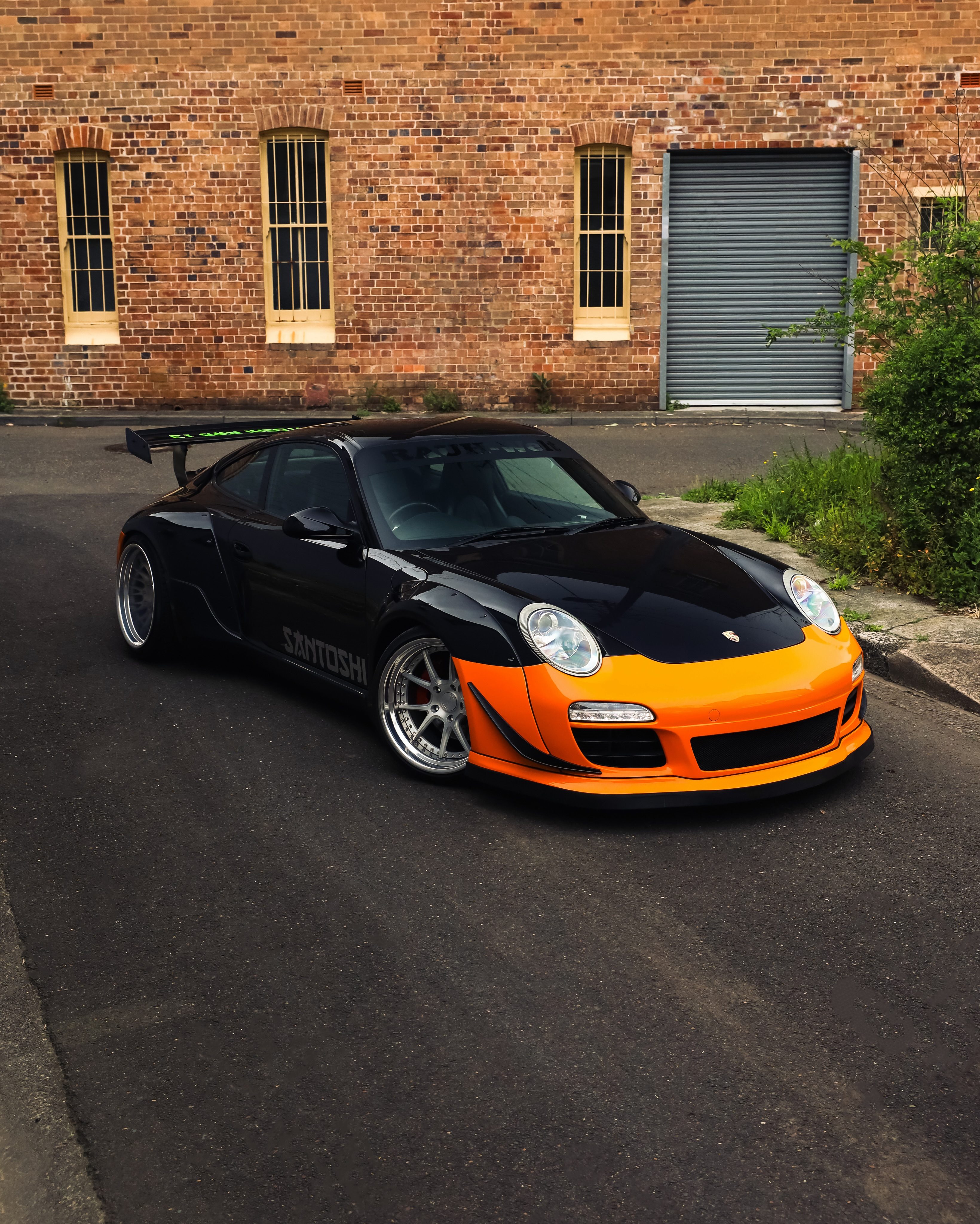 Black RWB widebody Porsche 911 with an orange front bumper, in a Sydney back lane against a brick wall