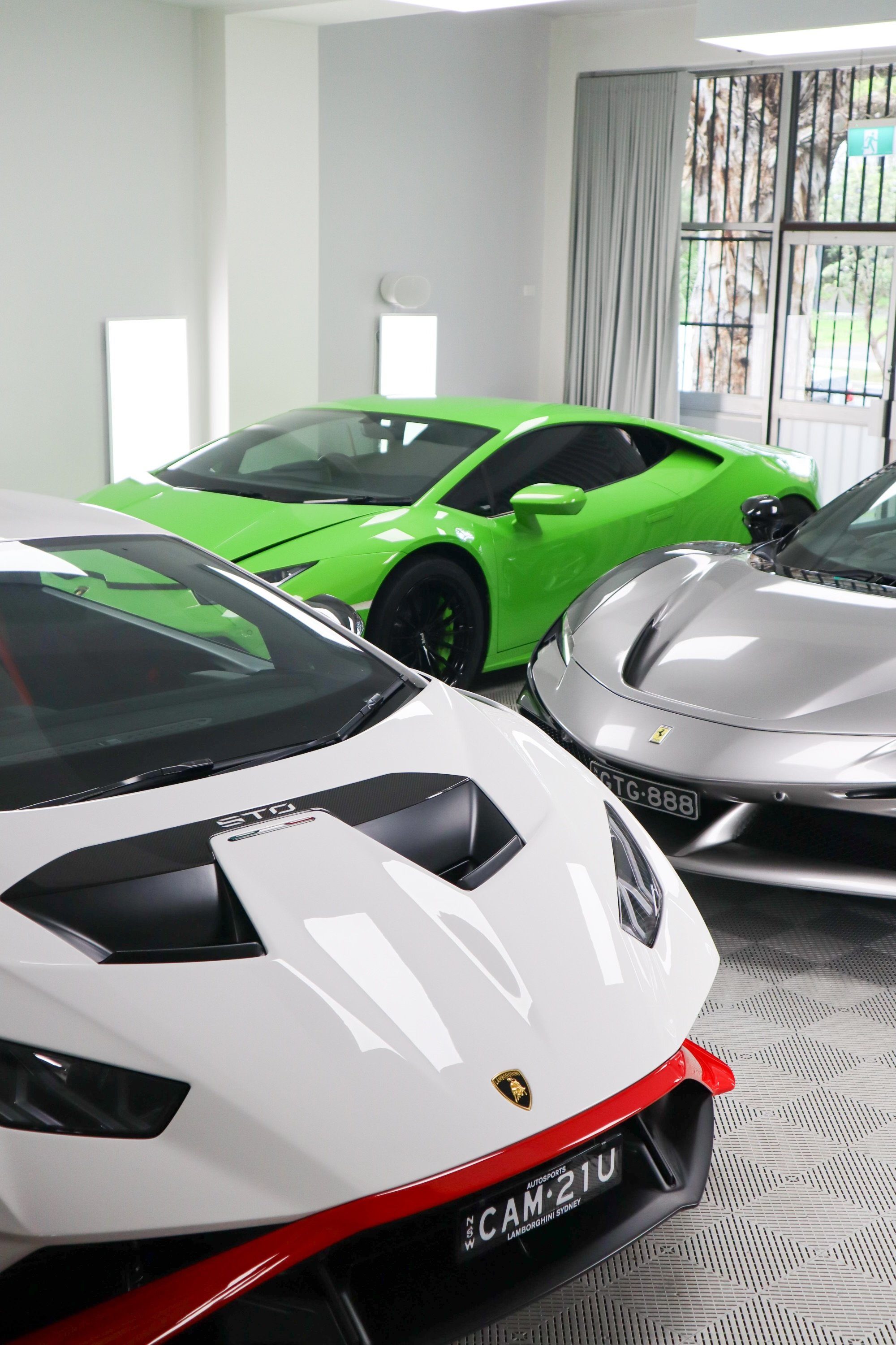 A white Lamborghini Huracan STO, a green Huracan and a silver Ferrari parked together in a light filled showroom